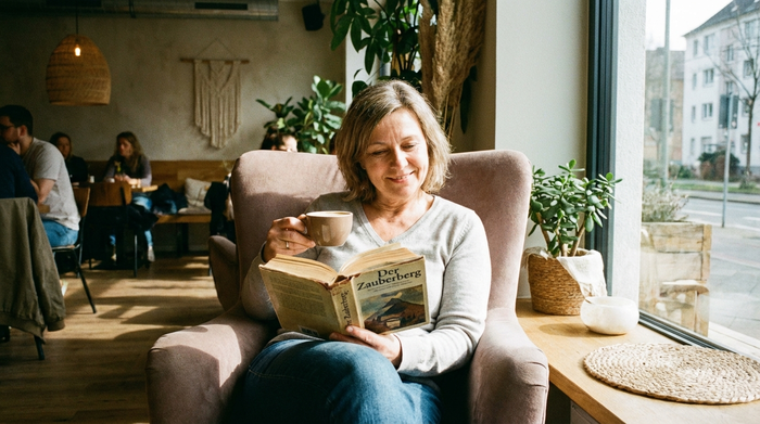 Eine entspannte Frau mittleren Alters trinkt in Ruhe einen Kaffee in einem modernen Café und liest ein Buch. Sie wirkt gelöst, erholt und glücklich. Weiches Licht, gemütliche Atmosphäre.