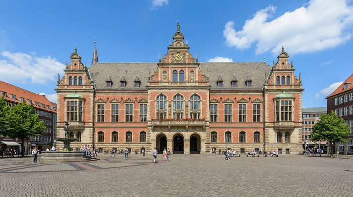 Ein gepflegtes, historisches Verwaltungsgebäude in einer deutschen Stadt an einem sonnigen Tag. Menschen spazieren entspannt über den Vorplatz. Klare, realistische Fotografie mit blauem Himmel.