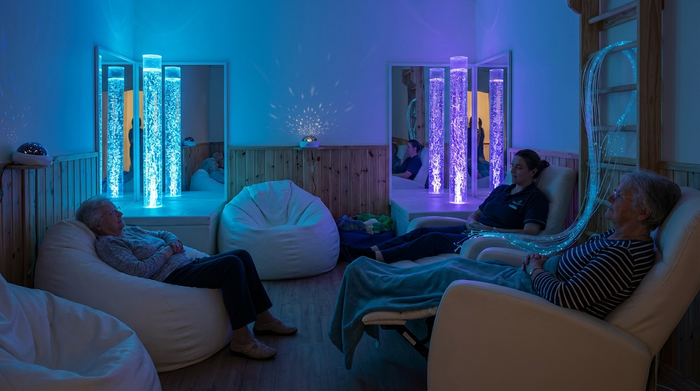 Ein gemütlicher Snoezelen-Raum mit gedimmtem, beruhigendem blauen Licht. Im Raum stehen sanft blubbernde, beleuchtete Wassersäulen und bequeme weiße Sitzsäcke. Entspannende, therapeutische Umgebung für Senioren, fotorealistisch.