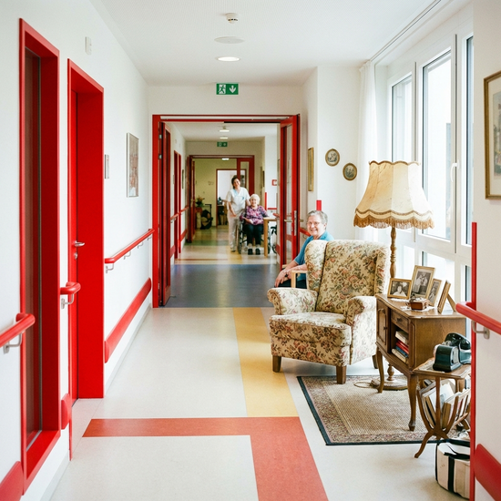 Ein heller, freundlicher Flur in einer Pflegeeinrichtung mit starken Farbkontrasten zur besseren Orientierung. Rote Handläufe heben sich deutlich von weißen Wänden ab. Eine gemütliche Erinnerungsinsel mit einem antiken Sessel und einer alten Stehlampe lädt zum Verweilen ein.