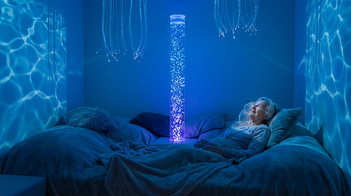 Ein beruhigender Snoezelraum mit sanftem, bläulichem Licht. Eine ältere Dame liegt entspannt auf einer bequemen Liegelandschaft und betrachtet fasziniert eine leuchtende, sprudelnde Wassersäule. Die Stimmung ist meditativ, entspannend und friedvoll.