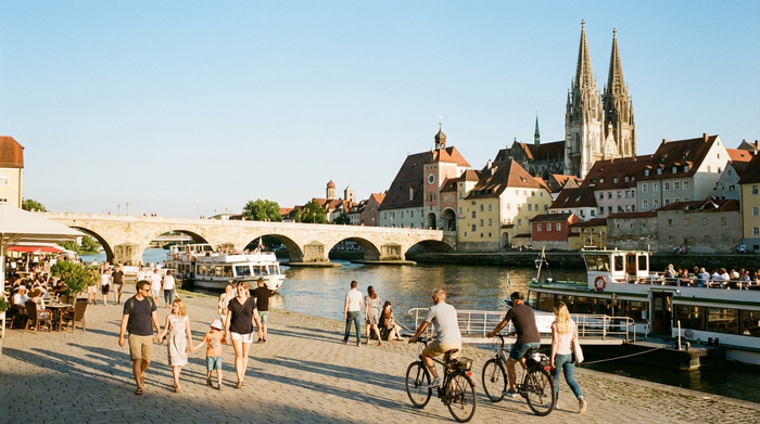 Blick auf die historische Altstadt von Regensburg mit der Steinernen Brücke im Hintergrund, im Vordergrund spazieren Menschen entspannt an der sonnigen Uferpromenade der Donau.