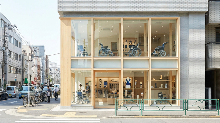 Die helle, einladende Außenfassade eines modernen Sanitätshauses an einer belebten Straße. Große Schaufenster zeigen dezente medizinische Hilfsmittel. Klares Wetter, realistische Szene ohne Schriftzüge.