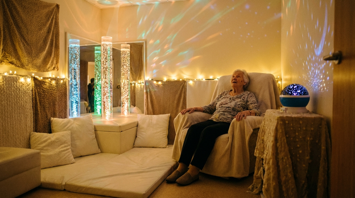 Ein gemütlicher Snoezelen-Raum mit gedimmtem, warmem Licht, sanft blubbernden Wassersäulen und weichen, weißen Sitzkissen. Eine ältere Dame sitzt entspannt in einem Sessel und betrachtet fasziniert die beruhigenden Lichtspiele in einer absolut friedlichen Umgebung.