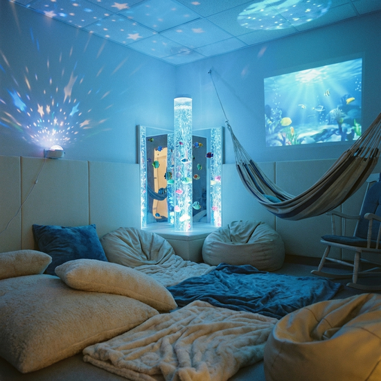 Ein beruhigender Snoezelen-Raum mit sanfter, bläulicher Beleuchtung und einer sanft blubbernden Wassersäule. Gemütliche, weiche Sitzkissen liegen auf dem Boden. Die Atmosphäre ist extrem entspannend und friedlich. Ohne Text.