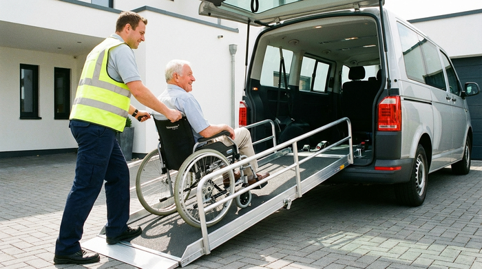 Ein Rollstuhlfahrer wird über eine sichere Rampe in einen geräumigen Transporter geschoben. Ein fürsorglicher Pfleger unterstützt ihn dabei. Helles Licht, saubere Umgebung, Fokus auf Sicherheit und Barrierefreiheit.