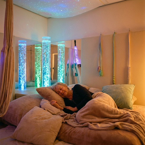 Ein gemütlicher Snoezelen-Raum mit gedimmtem, warmem Licht. Bequeme Sitzkissen und sanft leuchtende Wassersäulen schaffen eine beruhigende Atmosphäre. Eine ältere Dame entspannt sich sichtlich in der friedlichen Umgebung. Fotorealistisch, harmonisch.