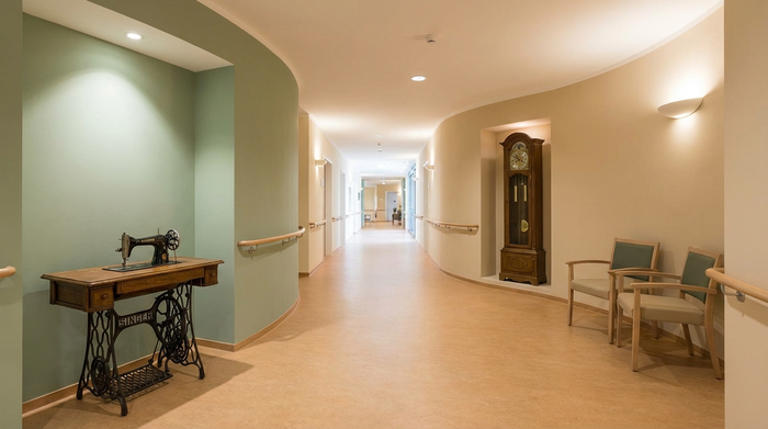 Ein heller, endloser Flur in einem modernen Pflegeheim, der als Rundweg angelegt ist. Sanfte Farbkontraste an den Wänden, historische Erinnerungsstücke wie eine alte Nähmaschine und eine Pendeluhr dienen als Orientierungshilfen. Ruhige, sichere Umgebung ohne Text.