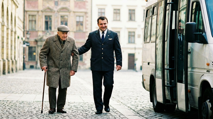 Ein fürsorglicher Fahrer reicht einem älteren Herrn mit Gehstock freundlich den Arm, während sie gemeinsam langsam auf einem gepflasterten Weg zu einem wartenden Kleinbus gehen. Im Hintergrund ist eine unscharfe Häuserfassade erkennbar. Helles Tageslicht.