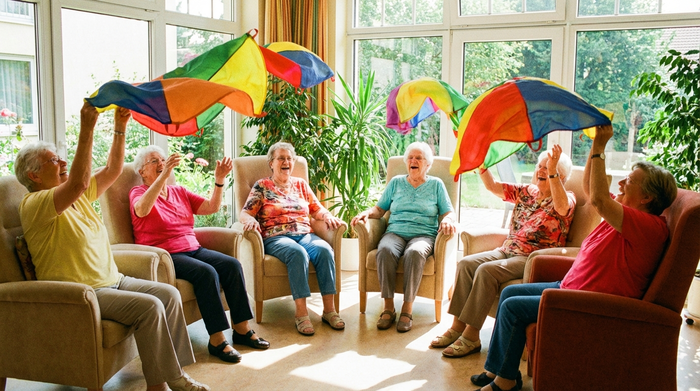 Senioren bei einer leichten Sitzgymnastik in einem lichtdurchfluteten Raum. Sie sitzen im Kreis auf bequemen Stühlen und heben bunte Schwungtücher in die Luft. Die Atmosphäre ist ausgelassen und aktiv. Viel Tageslicht und grüne Zimmerpflanzen im Hintergrund.