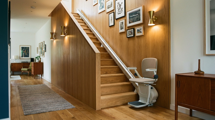 Ein moderner, unauffälliger Treppenlift ist an einer gepflegten Holztreppe in einem stilvollen Einfamilienhaus montiert. Warme Wandleuchten erhellen den Flur.