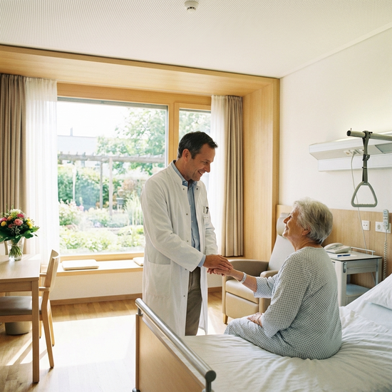 Ein freundlicher Arzt in weißem Kittel spricht beruhigend mit einer älteren Patientin in einem hellen, modernen Krankenhauszimmer. Tageslicht fällt durch das Fenster, positive und vertrauensvolle Atmosphäre, fotorealistisch.