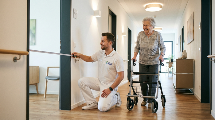 Ein professioneller, freundlicher Mitarbeiter eines Sanitätshauses in sauberer Arbeitskleidung misst mit einem Maßband die Türbreite in einem modernen Flur, während eine ältere Dame lächelnd zuschaut. Helle Beleuchtung, realistisch.