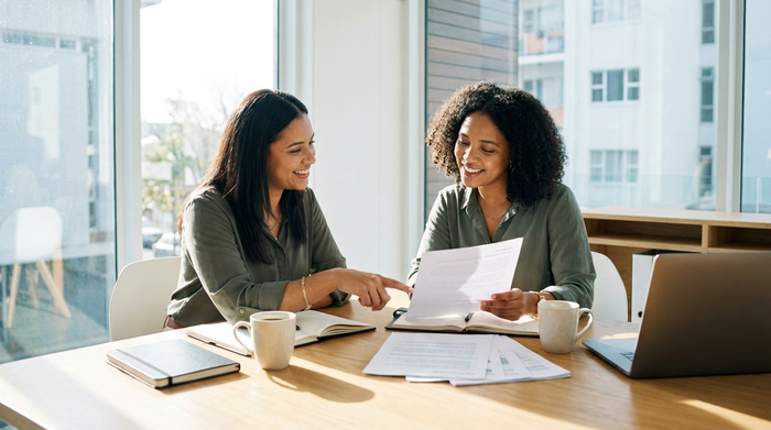 Zwei Frauen sitzen an einem Holztisch und besprechen Dokumente. Die Atmosphäre ist vertrauensvoll und professionell. Helles Büro, aufgeräumter Tisch mit Kaffeetassen. Fotorealistisch, ohne lesbaren Text.
