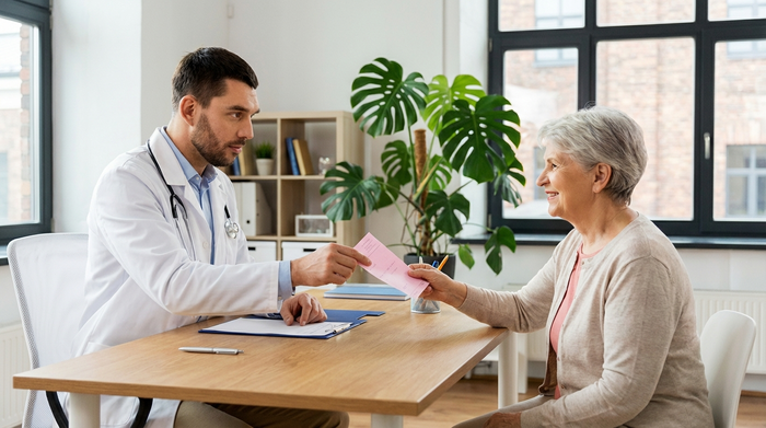 Ein fokussierter Arzt in einem weißen Kittel sitzt an einem aufgeräumten Holzschreibtisch und überreicht einer lächelnden Seniorin ein rosafarbenes Rezept. Helles, freundliches Praxiszimmer mit einer grünen Pflanze im Hintergrund.