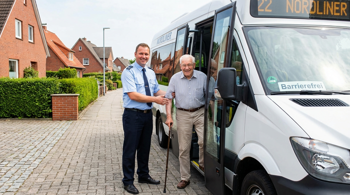 Ein freundlicher Busfahrer hilft einem älteren Herrn mit Gehstock lächelnd beim Einstieg in einen modernen, weißen Kleinbus. Helles Tageslicht, eine typische norddeutsche Wohnsiedlung mit Backsteinhäusern im Hintergrund.