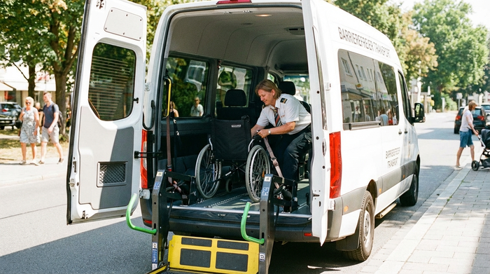 Eine ausgefahrene Hebebühne am Heck eines weißen Kleinbusses. Ein Fahrer sichert behutsam einen leeren Rollstuhl im Innenraum mit speziellen Gurten. Helle, freundliche Atmosphäre.