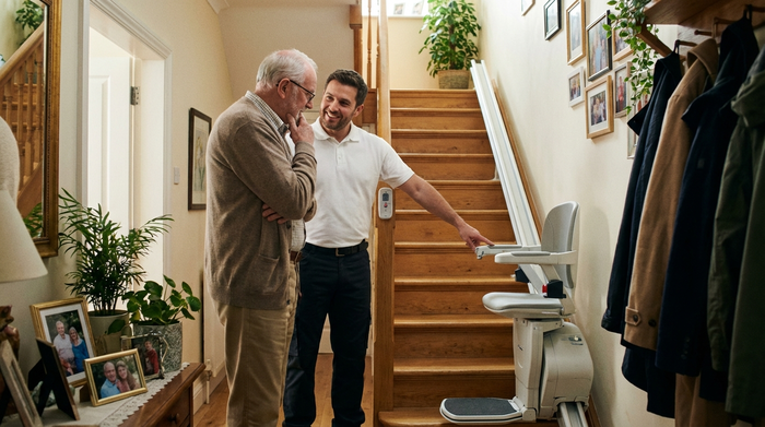 Ein freundlicher Techniker in sauberer Arbeitskleidung erklärt einem Senior im heimischen Flur die Funktion eines Treppenlifts. Helle Umgebung, vertrauensvolle Stimmung, realistische Alltagsszene ohne Text.
