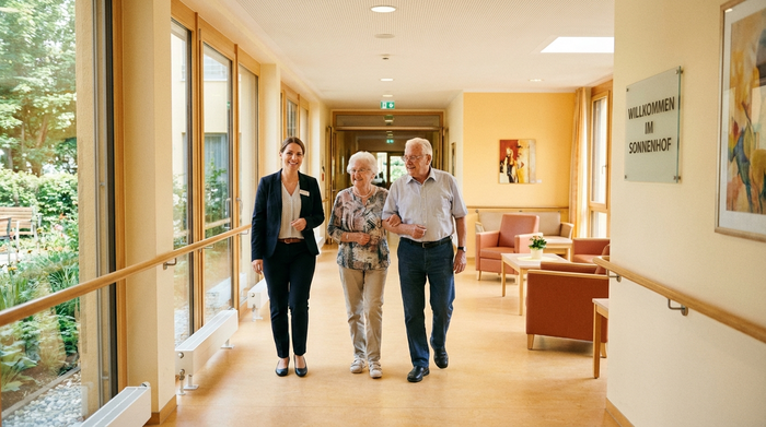 Eine freundliche Einrichtungsleiterin führt ein älteres Ehepaar durch einen breiten, hellen Flur eines modernen Pflegeheims. Der Flur hat warme Farben, Handläufe aus Holz und große Fenster. Einladende und sichere Umgebung.