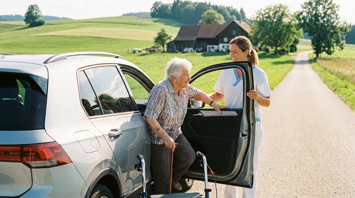 Eine Seniorin steigt mit Hilfe einer sympathischen Betreuungskraft sicher in ein modernes Auto ein. Ländliche Umgebung im Hintergrund, sonniger Tag, Fokus auf Mobilität.