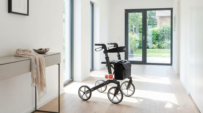 Ein eleganter, leichter Carbon-Rollator steht in einem hellen, modernen Flur eines Wohnhauses. Die Szene ist aufgeräumt und strahlt Mobilität und Sicherheit aus.