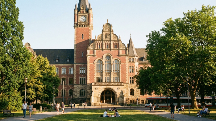 Ein historisches, gepflegtes Verwaltungsgebäude im Ruhrgebiet an einem sonnigen Tag, umgeben von grünen Bäumen und spazierenden Menschen. Die Architektur strahlt Beständigkeit und öffentliche Fürsorge aus.