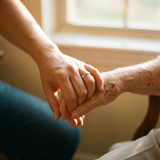 Eine Nahaufnahme von Händen, die sanft übereinandergelegt sind. Eine jüngere Hand hält tröstend die Hand einer älteren Person. Warme Beleuchtung, Fokus auf Geborgenheit.