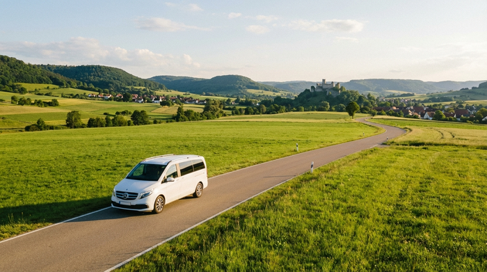 Ein moderner weißer Kleinbus fährt auf einer malerischen Landstraße entlang grüner Wiesen am Fuße der Schwäbischen Alb. Sonniges Wetter, weite Landschaft, sichere und ruhige Fahrt.