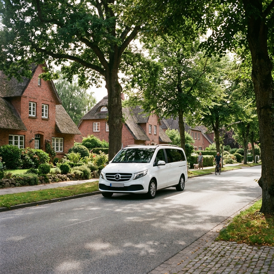 Ein moderner, weißer Kleinbus fährt durch eine gepflegte norddeutsche Vorstadtstraße mit grünen Bäumen an einem sonnigen Tag. Ruhige, friedliche Atmosphäre, realistische Darstellung ohne Text.