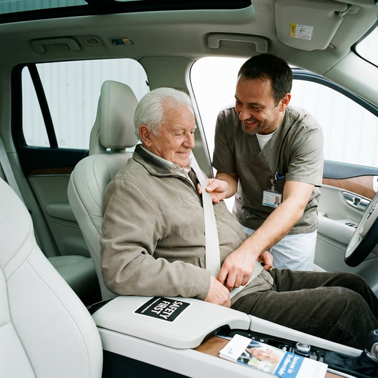 Ein freundlicher Pfleger befestigt behutsam einen Sicherheitsgurt an einem bequemen Autositz, auf dem ein zufriedener Senior sitzt. Helles Interieur, Fokus auf Sicherheit und Fürsorge.