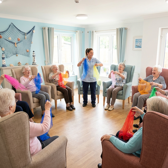 Senioren sitzen im Kreis in bequemen Sesseln und machen leichte Gymnastikübungen mit bunten Tüchern. Eine engagierte Betreuungskraft macht die Bewegungen vor. Helles, freundliches Zimmer mit maritimer Dekoration, fröhliche Stimmung.