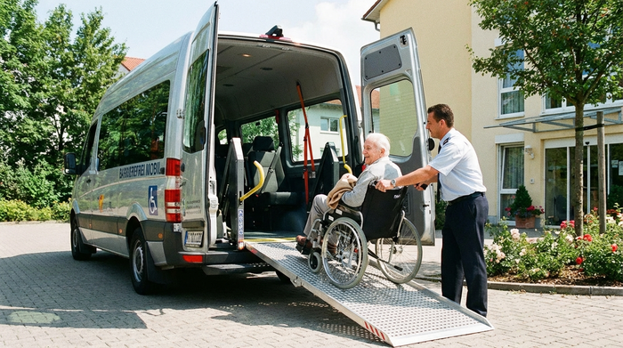 Ein geräumiger Kleinbus mit ausgefahrener, stabiler Metallrampe am Heck. Ein fürsorglicher Fahrer schiebt behutsam einen älteren Herrn im Rollstuhl in das helle Fahrzeuginnere. Fokus auf Barrierefreiheit und professionelle Ausstattung, sonniger Tag.