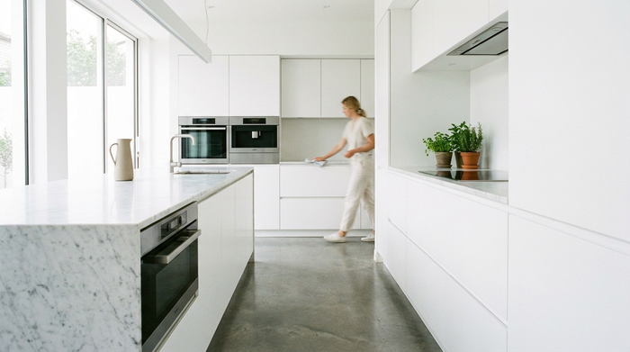 Eine saubere, moderne Küche. Im Hintergrund wischt eine Person unauffällig die Arbeitsfläche ab. Helles Tageslicht, aufgeräumte und hygienische Umgebung.