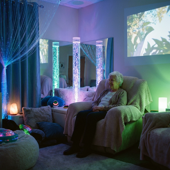 Ein gemütlicher Snoezelen-Raum mit gedimmtem, farbigem Licht. Eine ältere Person sitzt entspannt in einem bequemen Sessel und betrachtet sanft leuchtende Wassersäulen. Ruhige, therapeutische Atmosphäre, realistische Darstellung, ohne Text.