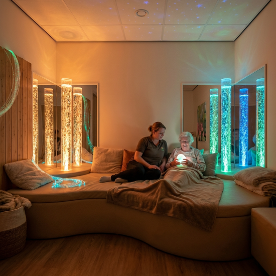 Ein gemütlicher Snoezelen-Raum mit gedimmtem, warmem Licht. Eine bequeme Liegelandschaft und sanft leuchtende Wassersäulen schaffen eine beruhigende Atmosphäre. Eine Pflegekraft sitzt entspannt neben einer älteren Dame.
