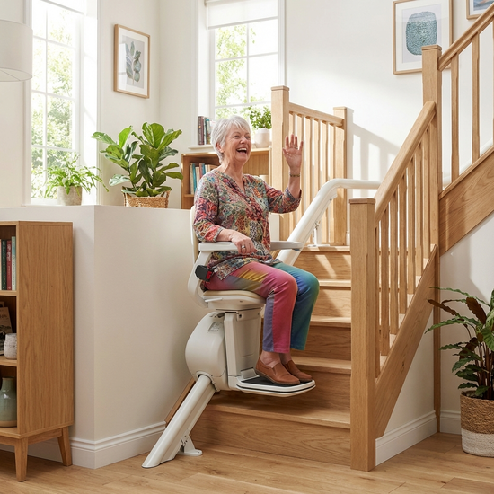 Eine fröhliche ältere Dame sitzt entspannt in einem modernen Treppenlift und fährt eine helle Holztreppe in ihrem gepflegten Zuhause hinauf. Sie lächelt sicher und zufrieden. Helle, einladende Wohnatmosphäre.