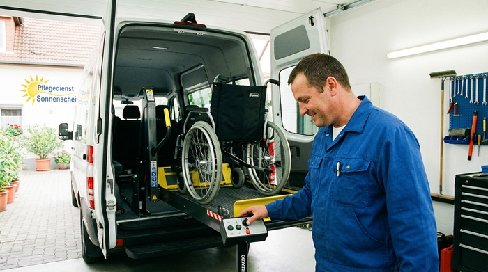 Ein freundlicher Pfleger bedient konzentriert eine hydraulische Hebebühne am Heck eines weißen Transporters, um einen leeren Rollstuhl sicher in das Fahrzeug zu heben. Gut beleuchtete, saubere Umgebung.