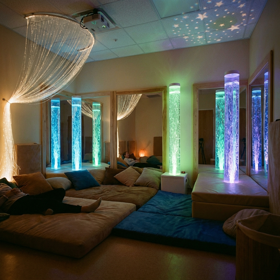 Ein gemütlich eingerichteter Snoezelen-Raum mit gedimmtem Licht, sanft leuchtenden Wassersäulen und bequemen, weichen Liegematten. Eine entspannende und beruhigende Atmosphäre.