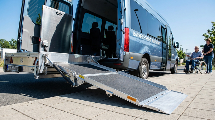 Die geöffnete Hecktür eines modernen Transportbusses mit einer ausgefahrenen, stabilen Metallrampe für Rollstühle. Ein gepflegter Gehweg im Vordergrund. Klarer Fokus auf die sichere Technik des Fahrzeugs, strahlender Sonnenschein.