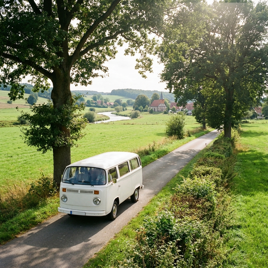 Ein weißer Kleinbus fährt an einem sonnigen Tag auf einer idyllischen Landstraße zwischen weiten, grünen Feldern. Fotorealistisch, ländliche niedersächsische Landschaft, ruhige Stimmung, ohne Beschriftungen.