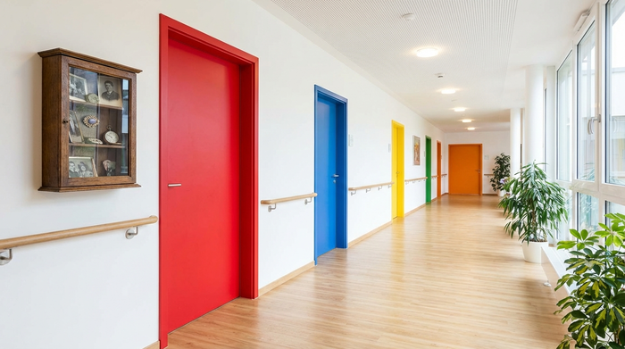 Ein heller, breiter Flur in einer modernen Senioreneinrichtung. Die Türen sind in kräftigen, unterschiedlichen Farben gestrichen, um die Orientierung zu erleichtern. Neben einer leuchtend roten Tür hängt ein kleiner Holzkasten mit persönlichen, antiken Erinnerungsstücken. Sauber, einladend, fotorealistisch und ohne Schilder oder Text.