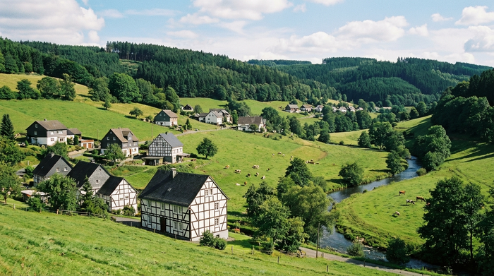 Ein malerischer Blick auf die hügelige Landschaft des Siegerlandes im Sommer, mit verstreuten Einfamilienhäusern an grünen Hängen. Fachwerkhäuser und dichte Wälder im Hintergrund, klare, realistische Fotografie ohne Text.