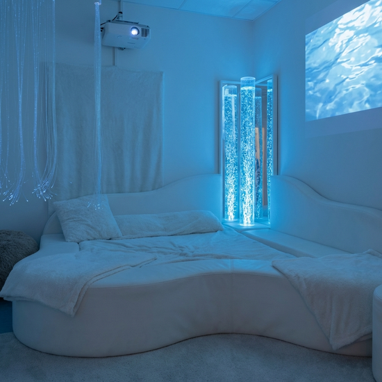 Ein beruhigender Snoezelen-Raum in sanftes, blaues Licht getaucht. Eine bequeme, weiße Liegelandschaft, daneben eine sanft blubbernde, beleuchtete Wassersäule. Eine friedliche Atmosphäre ohne Personen.