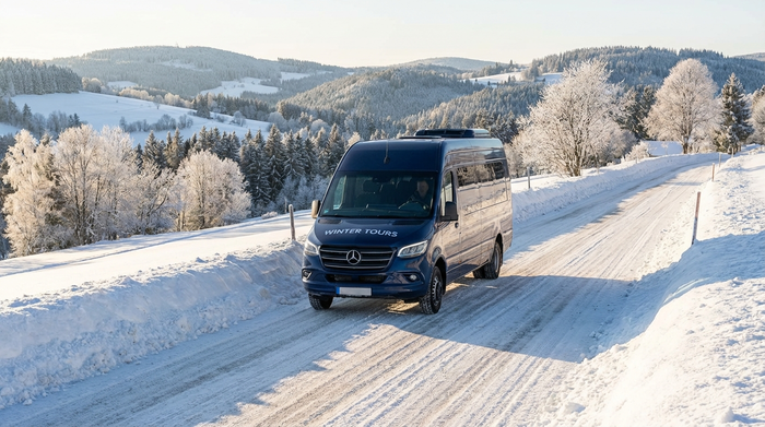Ein gepflegter Transportbus fährt vorsichtig eine leicht verschneite, gut geräumte Straße in einer hügeligen Winterlandschaft hinauf. Die Wintersonne strahlt auf den glitzernden Schnee am Straßenrand. Sichere Fahrweise im Winter.