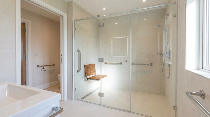 Ein modernes, barrierefreies Badezimmer mit einer großzügigen, bodengleichen Dusche aus Glas. Helle Fliesen, ein eleganter Duschklappsitz aus Holz und verchromte Haltegriffe. Sauber, luxuriös und sicher.
