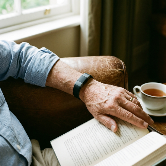 Ein moderner, unauffälliger Hausnotruf-Knopf am Handgelenk eines älteren Mannes, der entspannt ein Buch liest. Fokus auf das Handgelenk und das Gefühl von Sicherheit.