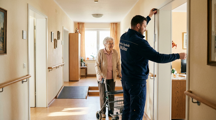 Ein professioneller Reha-Techniker in gepflegter, dunkelblauer Arbeitskleidung misst mit einem Maßband die Türbreite in einem gemütlichen, barrierefreien Flur einer Seniorenwohnung. Eine ältere Dame schaut freundlich zu.