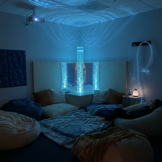 Ein gemütlich eingerichteter Snoezelen-Raum mit gedimmtem, beruhigendem blauem Licht. Eine leuchtende Wassersäule mit sanft aufsteigenden Blasen steht im Zentrum, umgeben von weichen Sitzkissen und entspannender Atmosphäre.