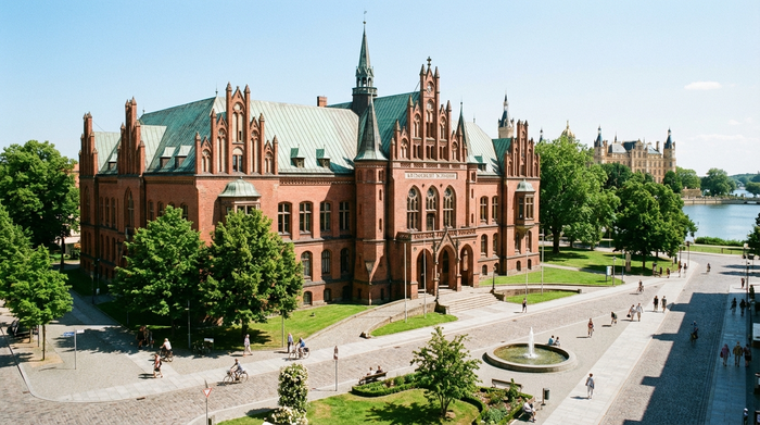 Ein gepflegtes, historisches Amtsgebäude in Schwerin mit roter Backsteinfassade an einem hellen Tag, umgeben von grünen Bäumen und sauberen Gehwegen.