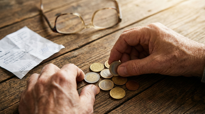 Eine Nahaufnahme von Händen, die ein paar Euro-Münzen auf einem rustikalen Holztisch zählen. Eine unscharfe Brille liegt im Hintergrund. Helle Beleuchtung, symbolisiert die gesetzliche Zuzahlung.